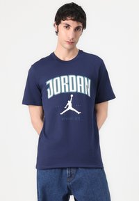 Mørkeblå bomulds t-shirt med "JORDAN" trykt i teal, med et hvidt silhuetlogo nedenunder. Korte ærmer og rund hals.