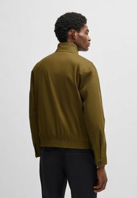Olive green bomber jacket with a stand-up collar, elastic waistband, and buttoned cuffs. Smooth, lightweight fabric with a minimalist design.