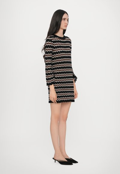 Robe mini en tricot noire avec un motif ondulé blanc et beige. Manches longues et ourlet en dentelle. Le modèle porte des chaussures à talons ouverts noires.
