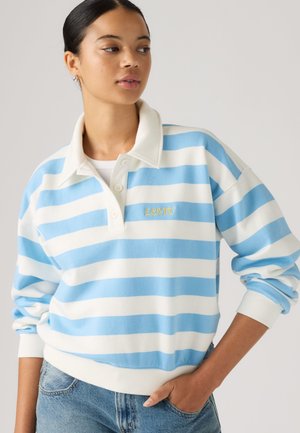 Femme portant un polo à manches longues rayé blanc et bleu avec le logo "Levi's", debout avec une main dans la poche d'un jean bleu.