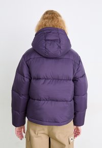 Vijolična napihljiva jakna s kapuco, ki vključuje prešito zasnovo, gladko teksturo in elastične manšete. Uporabljena je z rjavimi cargo hlačami.