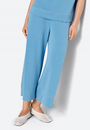 Nő világoskék, széles szárú, rövidített nadrágot és fehér hálós, felhúzható cipőt visel, fehér háttér előtt áll.