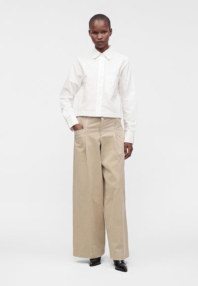 Femme portant une chemise blanche boutonnée, rentrée dans un pantalon beige à jambes larges, les mains dans les poches, debout devant un fond uni.