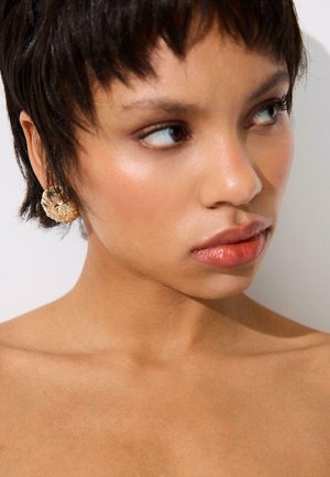 Woman with short dark hair wearing a large gold floral earring, looking to the side with a neutral expression against a plain background.