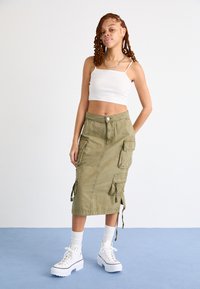 Jupe cargo vert olive avec poches latérales et liens, associée à un haut court blanc et des baskets blanches épaisses à semelles compensées.