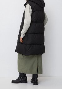 Svart pufferväst med huva, som har quiltade sektioner och en åtsittande midja. Kombineras med en lång olivgrön kjol och svarta stövletter.
