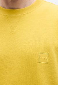 Κίτρινο φούτερ με υφή, με στρογγυλή λαιμόκοψη και κεντημένο τετράγωνο λογότυπο στο στήθος. Απλός σχεδιασμός χωρίς σχέδια.