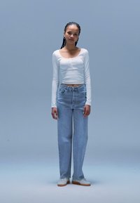 Top branco cropped de manga comprida, jeans azul-claro de perna reta e sapatilhas brancas com sola em borracha, sobre um fundo azul suave.