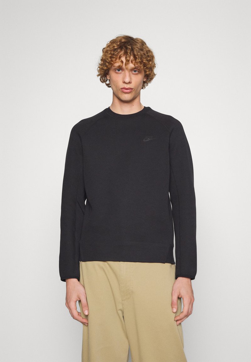 Jeune homme aux cheveux bouclés portant un sweat à capuche noir Nike et un pantalon beige, se tenant devant un fond blanc uni.