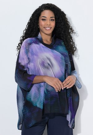 Femme aux longs cheveux bouclés portant un haut ample à motifs violets et bleus, souriante avec les mains doucement jointes devant sur un fond blanc.