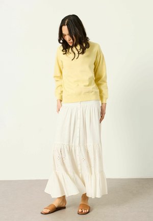 Frau steht drinnen, trägt einen gelben Pullover, langen weißen Rock mit Knöpfen und braune gewebte Slipper-Sandalen, schaut nach unten.