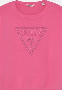 Rosa tröja med en upphöjd, präglad triangulär logotyp med rhinestones och texten "GUESS U.S.A". Ribbad krage och manschetter.