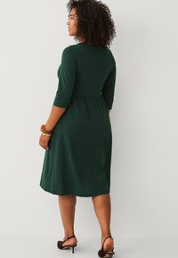 Grøn kjole i knælængde med 3/4-ærmer og en tætsiddende talje. Lavet af et blødt materiale, med et enkelt design og minimal dekoration.