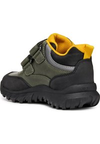 Sneaker verde con suola in gomma nera, dettagli grigi e fodera in rete gialla. Presenta due cinturini regolabili in velcro e una linguetta sul retro per facilitare l'uso.