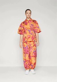 Sieviete stāv ar spilgti oranžu krekla un biksēm, kurām ir lieli rozā un purpursarkanie tropisko lapu raksti, kā arī baltām kedām.