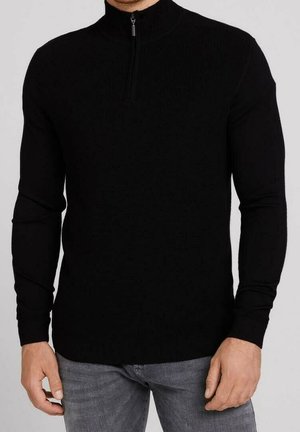 Homme portant un pull noir à manches longues avec fermeture éclair à moitié et un jean gris, debout devant un fond clair uni.