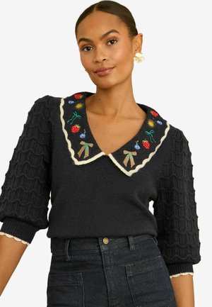 Žena v čiernom pletenom svetri s nadýchanými rukávmi a vyšívaným golierom s motívmi jahôd, mašlí a kvetov, v kombinácii s tmavými džínsy.