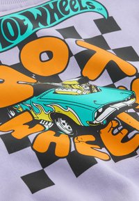 Egy világoslila póló, amelyen türkiz színű autó grafikája látható sárga lángdíszítéssel, és élénk narancssárga szöveg fekete kockás háttérrel.