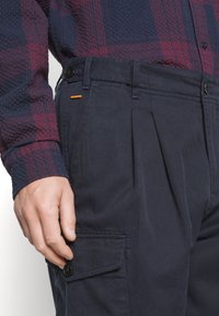 Pantaloni cargo navy con pieghe, dotati di una tasca con bottone sul lato e un'etichetta arancione in evidenza in vita. Indossati con una camicia a scacchi.