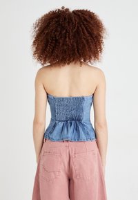 Pessoa com cabelo encaracolado usa um top azul sem mangas e calças largas cor-de-rosa, de costas para a câmara, contra um fundo branco simples.