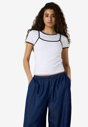 Young woman with long curly hair wearing a white fitted T-shirt with black trim and loose dark blue pants with hands in pockets.