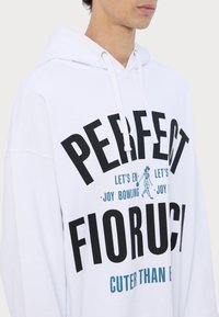 Hvid hoodie med sort og teal grafisk tekst. Har snoretræk i hætten og en afslappet pasform. Teksten inkluderer "PERFECT" og "FIORUCI."