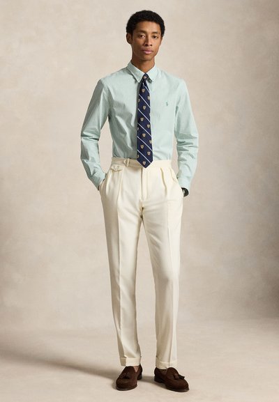 Homme portant une chemise rayée vert clair, une cravate à blason marine, un pantalon plissé crème taille haute et des mocassins à glands marron, debout les mains dans les poches.