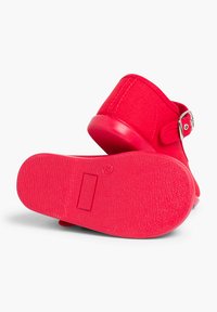Sandal rojo con suela de goma texturizada, parte superior de tela y detalle de hebilla metálica. La forma general es de punta abierta y sin talón.