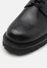 Chaussure à lacets en cuir noir avec bout rond et semelle épaisse en caoutchouc, présentée en gros plan sur un fond blanc uni.