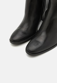 Botas de cuero negro hasta el tobillo con punta afilada, textura suave y un brillo sutil. Cuentan con un cierre lateral y un tacón bajo y duradero.