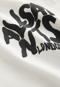 Czarne pogrubione litery tworzące "ALSA NYS LONDON" na białej tkaninie, pokazane z bliska z lekkimi fałdami i cieniami.