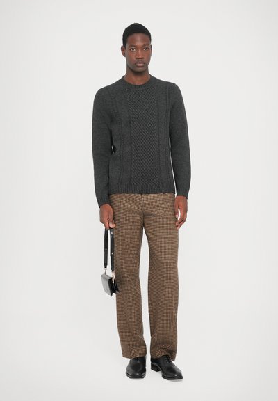 Pull en tricot gris foncé avec des motifs texturés, associé à un pantalon à carreaux marron. Le modèle tient un petit sac noir et gris. Chaussures noires.