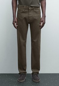 Pantalons en coton marron à coupe droite, avec une taille standard, des poches avant et des détails de couture subtils. Associés à des chaussures grises.