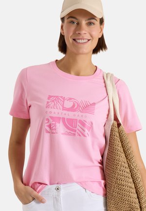 Femme portant une casquette beige, un t-shirt rose « Coastal Garden », un pantalon blanc et portant un grand sac tissé sur son épaule, souriante.