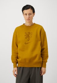 Kollane hetshert, mis on valmistatud pehmest kangast, omades lõviga graafikat ja Nike logo punktide disainis, ribidega varrukate ja alumise äärega.
