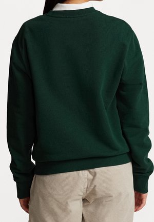 Persoon die een donkergroene trui en lichtbeige corduroy broek draagt, met de rug naar de camera toe staand tegen een effen achtergrond.