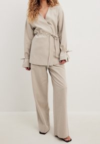 Kvinna iförd en beige omslagsjacka med midje- och ärmband, matchande vida byxor och svarta sandaler med öppen tå framför en neutral bakgrund.