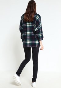 Flanelowa koszula w ciemnozielonym kolorze w kratę w odcieniach kremowym i fioletowym. Charakteryzuje się luźnym krojem, opuszczonymi ramionami i jest zestawiona z czarnymi obcisłymi dżinsami.
