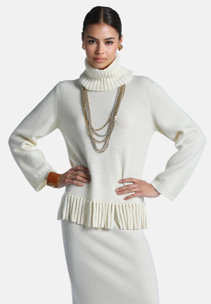 Femme aux cheveux foncés portant un pull à col roulé blanc à manches longues avec un ourlet à franges, une jupe assortie, des chaînes dorées superposées, des boucles d'oreilles dorées et un bracelet manchette marron.