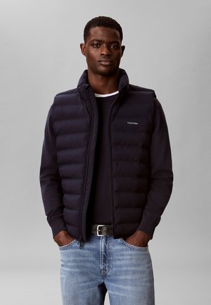 Uomo che indossa un gilet trapuntato nero sopra una camicia nera a maniche lunghe, jeans blu e cintura nera, in piedi con le mani in tasca su sfondo grigio.