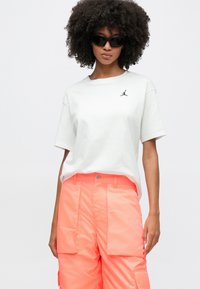 Göndör hajú nő fekete napszemüvegben, fehér pólóban, amelyen ugró ember logó látható, és élénk korallszínű cargo nadrágban fehér háttér előtt.