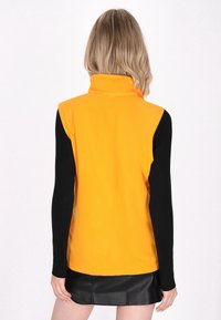 Gilet in pile giallo con collo alto, indossato sopra maniche lunghe nere, presenta una texture liscia e una vestibilità su misura. Vista posteriore mostrata.