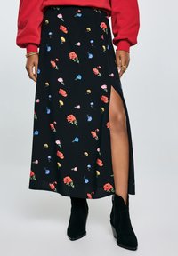 Kvinde iført en rød sweatshirt og en sort blomsterkjørt med en sideslids, parret med sorte ankelstøvler, mod en neutral baggrund.