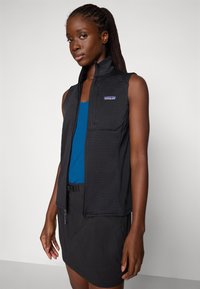 Kobieta z warkoczem, ubrana w czarną kamizelkę na zamek bez rękawów na niebieskim topie i czarnej spódnicy, stojąca na jednolitym tle.