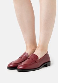Ein Paar glatte, burgunderfarbene Leder Slipper, die barfuß getragen werden, mit niedrigen schwarzen Sohlen und einem klassischen Penny-Strap-Design.
