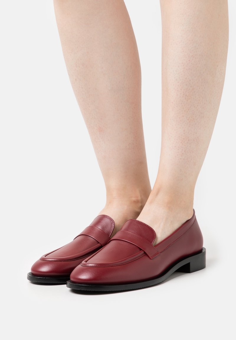 Ein Paar glatte, burgunderfarbene Leder Slipper, die barfuß getragen werden, mit niedrigen schwarzen Sohlen und einem klassischen Penny-Strap-Design.