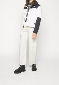 Biała i czarna ocieplana kurtka z wysokim kołnierzem, połączona z beżowymi spodniami cargo i czarnymi, masywnymi butami. Teksturowany materiał z widocznymi kieszeniami.