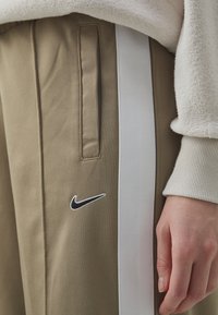 Béžové Nike tepláky s bílým bočním pruhem. Mezi vlastnosti patří přední kapsy a černě vyšitá logo na stehně. Měkká textura látky.