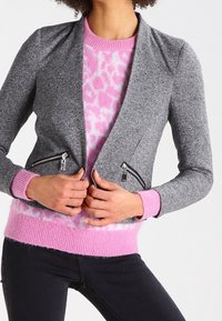 Šedý blazer s přiléhavým střihem, dvěma zipovými kapsami, klopovým límcem a pod ním měkký růžový svetr se vzorem leopardí kůže.
