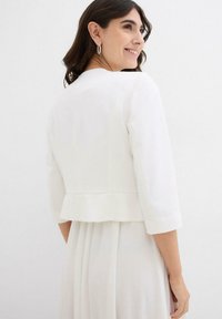 Giacca bianca accorciata con maniche a tre quarti, caratterizzata da un orlo peplum. Tessuto liscio con dettagli minimi, indossata sopra un abito coordinato.